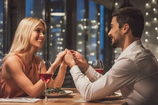 Na obrazie widać uśmiechniętą parę siedzącą przy stole w restauracji, trzymającą się za ręce nad stołem. Przed nimi znajdują się dwa kieliszki czerwonego wina oraz talerze z jedzeniem. W tle widoczna jest szyba, przez którą widać światła miasta