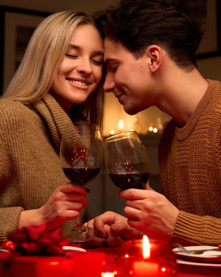 Na obrazku widzimy uśmiechniętą parę zakochanych ludzi, którzy siedzą blisko siebie, każda trzyma kieliszek czerwonego wina. Wygląda na to, że wznoszą toast lub dzielą się chwilą, z twarzami blisko siebie