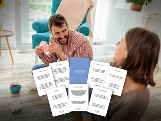 Grafika przedstawia talie kart gry na prezent Emocje Nasze Rozmowy na tle pary grającej razem w karty mężczyzna trzyma talie i uśmiecha się w kierunku kobiety.