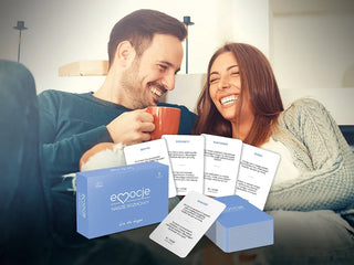 Grafika przedstawia przód niebieskiego opakowania gry na prezent dla par Emocje Nasze Rozmowy oraz talię do gry na tle uśmiechającego się mężczyzny trzymającego kubek i śmiejącej się koło niego kobiety siedzącej na sofie. 