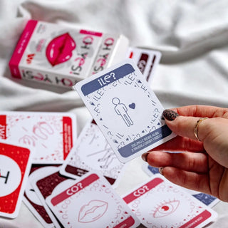 Zdjęcie przedstawia kobiecą dłoń trzymającą przykładową kartę z tali gry dla par oh yes pink, przedstawiająca liczbę rzeczy, które należy z siebie zdjąć. W tle opakowanie gry i pozostałe karty rozsypane obok.